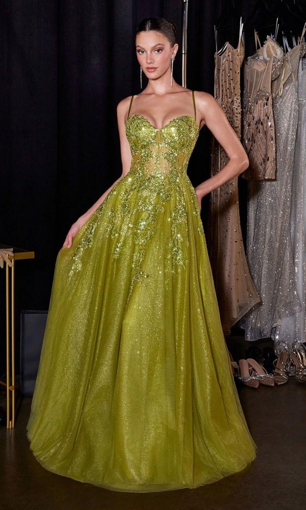 A woman stands in a dressing room with dresses behind her, wearing the Ladivine C177 long prom dress in olive, featuring a sparkly embellished bodice, spaghetti straps, and a full A-line skirt.